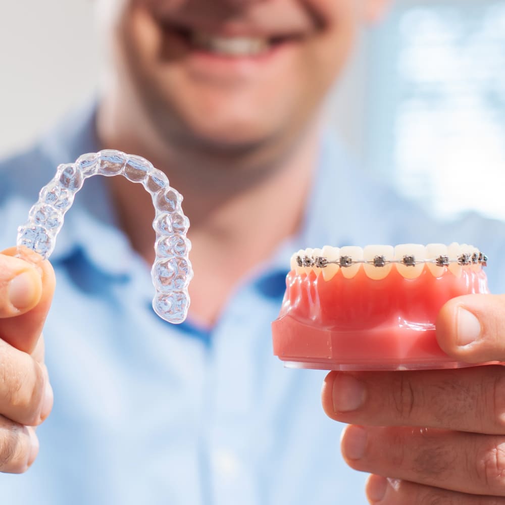 A dentist holds up an example of a mouth with Invisalign clear aligners in, and a set of teeth with traditional braces.