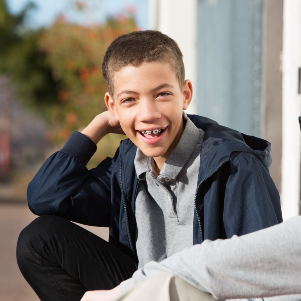 A young boy is crouching with his elbow on his knee. He is smiling, showing off his braces.