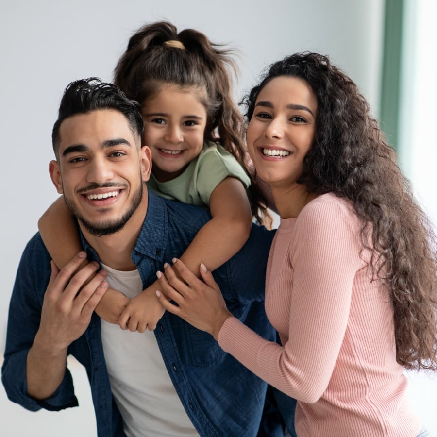 A family of three is smiling. The young girl is on her father's back.