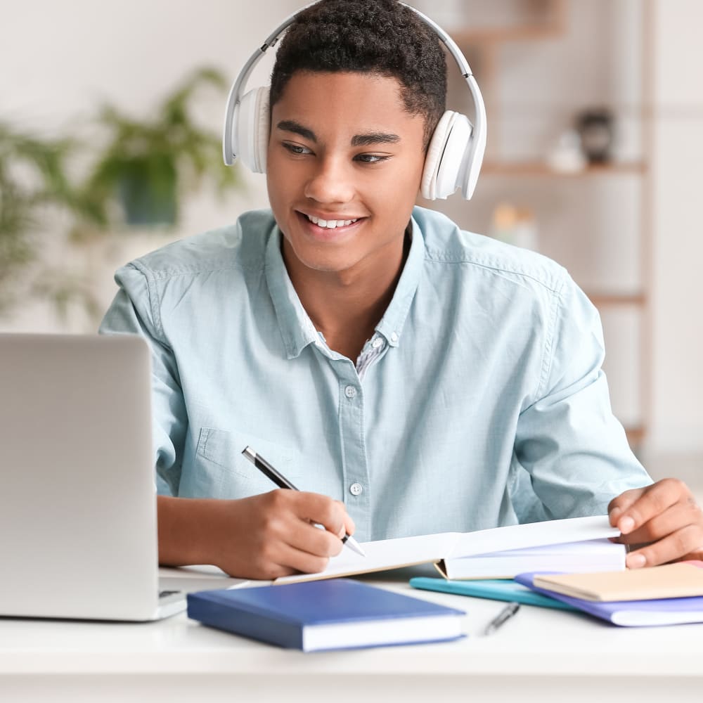 A young boy is wearing headphones, writing on paper, and looking at a laptop.